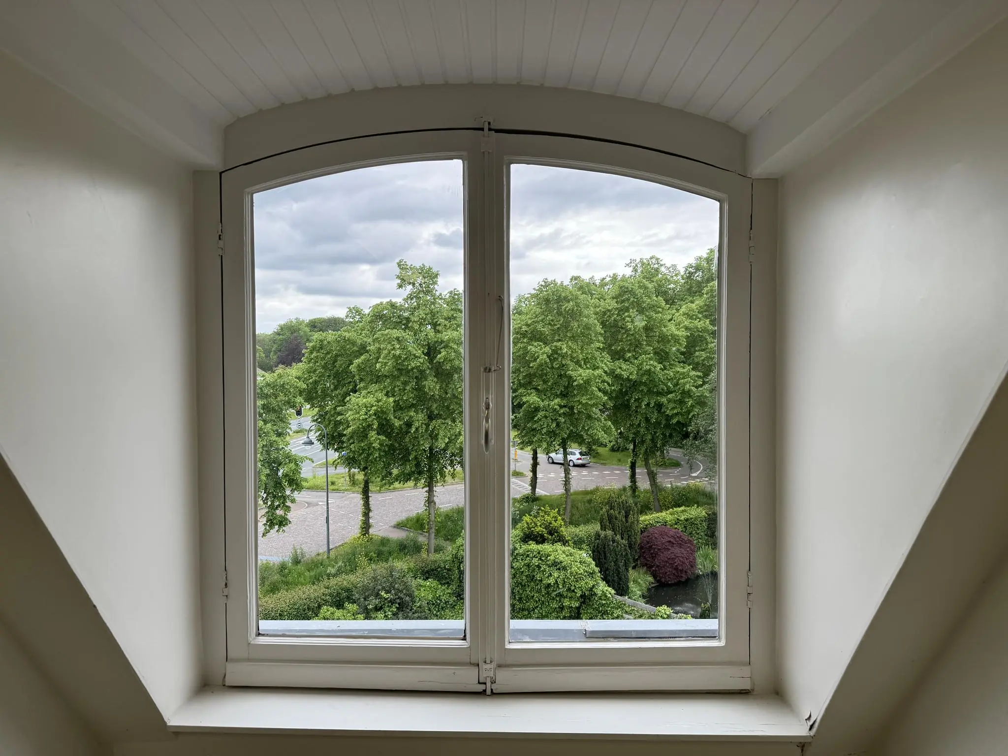 Uitzicht vanuit een zolderraam op de Dorpsstraat en Steenstraat met bomen, auto’s en groenvoorziening.